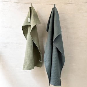 May include: Two waffle weave kitchen towels hanging on a clothesline. The towel on the left is sage green and the towel on the right is a darker green color called "storm".