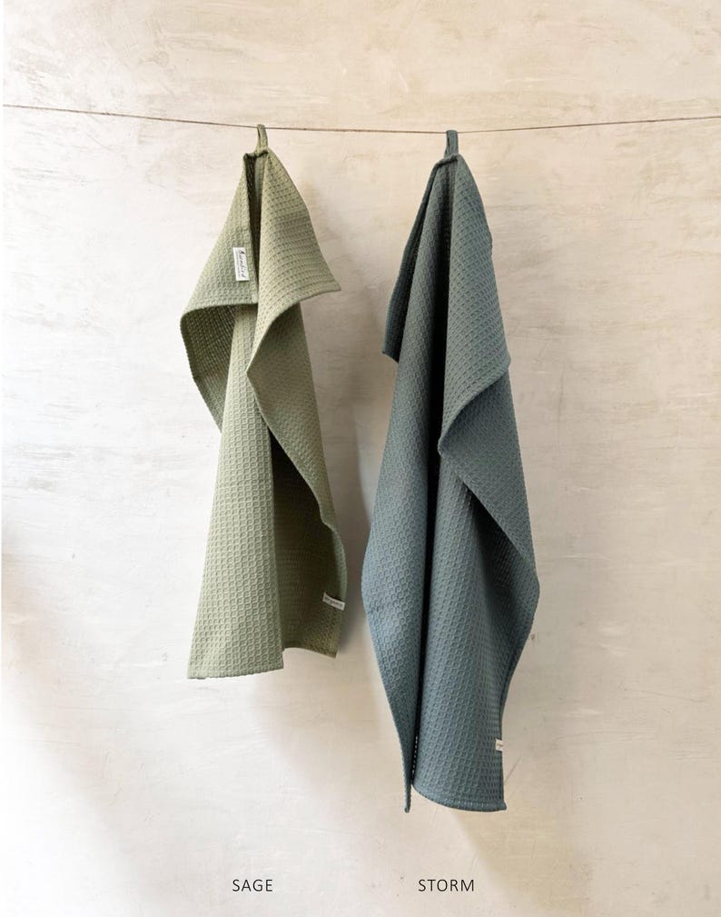 May include: Two waffle weave kitchen towels hanging on a clothesline. The towel on the left is sage green and the towel on the right is a darker green color called "storm".