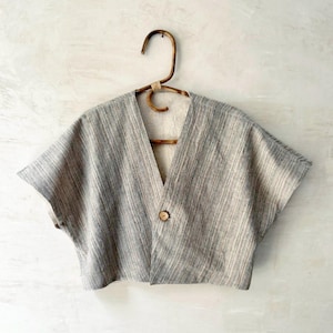 May include: A grey, short-sleeved top with a V-neck and a single wooden button closure. The top is made of a textured fabric and hangs on a wooden hanger.