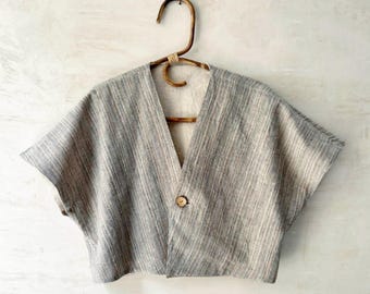 May include: A short-sleeved, cropped top in a light gray and white vertical striped pattern. The garment features a deep V-neckline, a single round wooden button closure at the center, and wide sleeves. It is displayed on a curved wooden hanger.