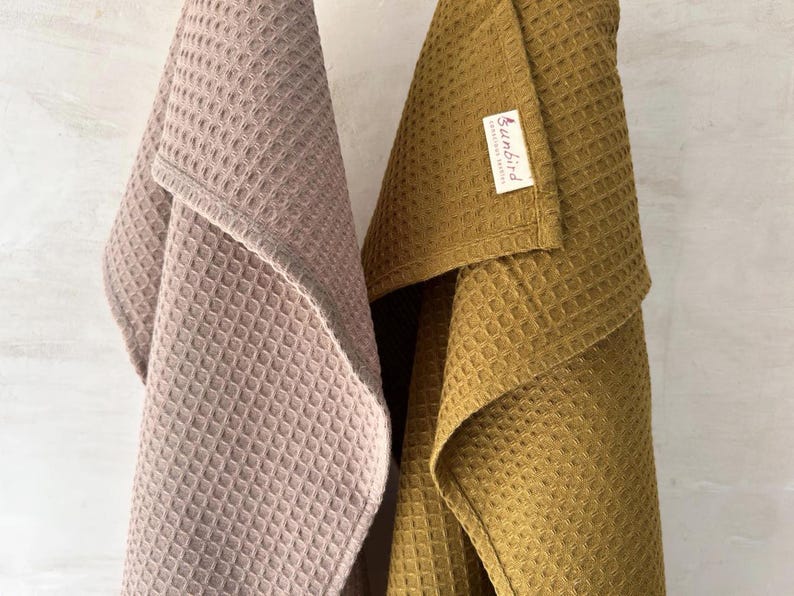 May include: Two waffle weave kitchen towels, one in a light brown color and one in a mustard yellow color. The yellow towel has a small label that says "Sunbird" on it.