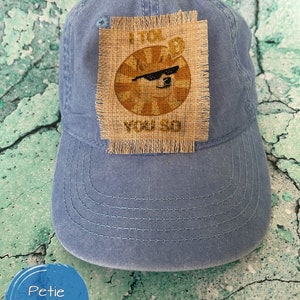 Puede incluir: Una gorra de béisbol de denim azul con un parche marrón que muestra un perro con gafas de sol y el texto "Te lo dije".