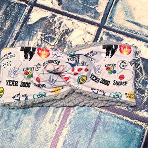 Puede incluir: Una diadema gris claro con un diseño de nudo retorcido. La diadema presenta un patrón con el texto "YEAR 3000" y otros gráficos. El interior está forrado con un material gris suave. La diadema está diseñada para la comodidad y el estilo.