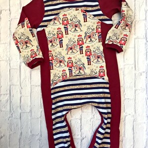 Puede incluir: Un mono de manga larga a rayas rojas, blancas y doradas con un estampado de Cascanueces en la parte delantera y las mangas.