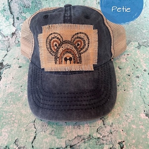 May include: A blue and beige trucker hat with a brown burlap patch featuring a cartoon mouse head with rainbow ears. The patch has frayed edges and the text "Petie Petie" is visible in the upper right corner of the image.