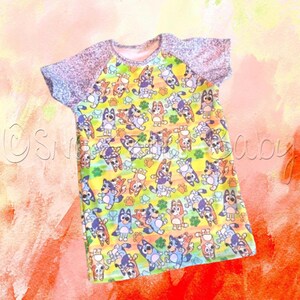 May include: A yellow, blue, and orange short-sleeved t-shirt with a repeating pattern of cartoon dogs. The shirt has a grey heather raglan sleeve.