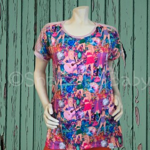 May include: A short-sleeved, round-neck t-shirt with a vibrant, all-over print featuring a repeating pattern of figures playing guitars and wearing colorful outfits. The shirt has a pink trim and is displayed on a mannequin.