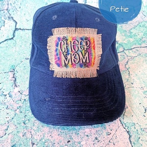 May include: A navy blue corduroy baseball cap with a frayed burlap patch that says "Cheer Mom" in rainbow letters with black leopard print accents.