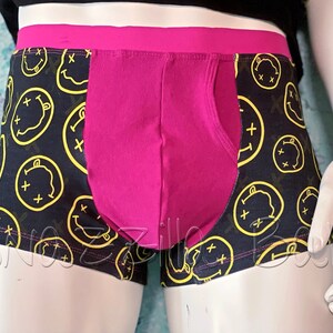 May include: Black boxer shorts with a pink waistband and a pink panel in the front. The boxer shorts have a repeating pattern of yellow smiley faces with an 'X' over the eyes.