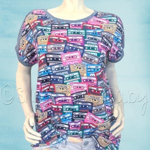 May include: A gray t-shirt with a print of colorful cassette tapes. The shirt has a round neckline and short sleeves.