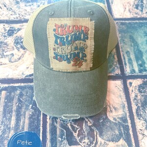 May include: A green and brown trucker hat with a distressed look. The hat has a square patch on the front with the words "TRUMP TRUMP TRUMP TRUMP 24" in red, white, and blue lettering on a burlap background.