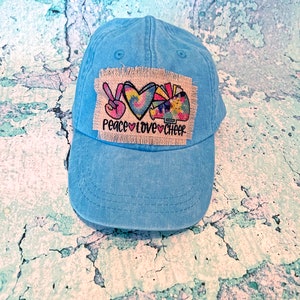 May include: A light blue denim baseball cap with a patch that says "Peace Love Cheer" and features a peace sign, a heart, and a cheerleader's pom-poms.