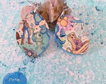 May include: A pair of dangle earrings with cartoon character designs. Each earring is an oval shape with a silver hook. One earring features characters from Toy Story and Monsters, Inc. The other earring features characters from Alice in Wonderland and Tangled.