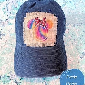 Peut inclure: Une casquette de baseball bleu foncé avec un patch en toile de jute effilochée représentant une tête de Mickey Mouse aux couleurs de l'arc-en-ciel avec un nœud rouge à pois blancs. Le texte "Petie Petie" est visible dans le coin inférieur droit de l'image.