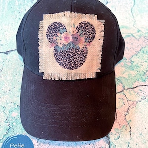 Peut inclure: Casquette de baseball noire avec un patch en toile de jute brune représentant une tête de Mickey Mouse à imprimé léopard avec des accents floraux.