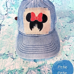 May include: A light blue denim baseball cap with a Minnie Mouse design. The design features a red bow and black mouse ears on a burlap patch. The cap has a curved brim and a brown adjustable strap.