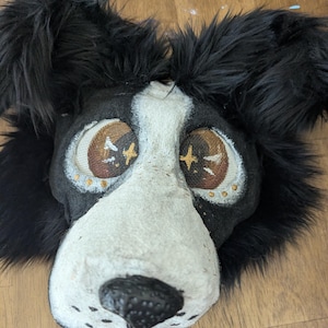 May include: A black and white animal mask featuring long black faux fur, a white muzzle, and a black nose. The eyes are large with brown irises, mesh screens, and painted star accents. The mask has a textured surface and upright ears.