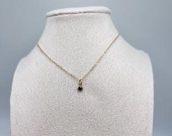 Può includere: Una delicata collana color oro con un piccolo pendente di colore scuro. La collana è esposta su un busto di manichino bianco su uno sfondo grigio chiaro. Il pendente è una piccola pietra rotonda di colore scuro.