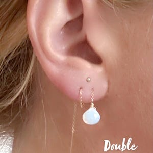 May include: A pair of gold double threaded earrings with a white opal gemstone hanging from each chain. The text "Double Threaded" is written below the earrings.