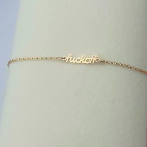 Op de afbeelding: Een delicate goudkleurige armband met het woord "fuckoff" in cursief geschreven. De armband bevindt zich op een lichte achtergrond. De ketting is dun en de letters zijn klein.