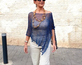 May include: A woman wearing a blue crochet poncho with a wooden bead necklace and white pants. She is wearing black sunglasses and walking on a brick walkway.