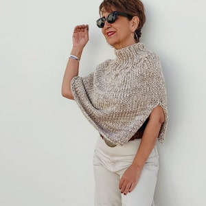 May include: A woman wearing a beige knitted poncho with a high neck and a brown belt. She is wearing white pants and black sunglasses.