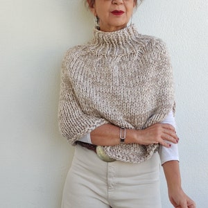 May include: A beige knitted poncho with a high neck and a subtle speckled pattern. The poncho is worn over a white shirt and beige pants.