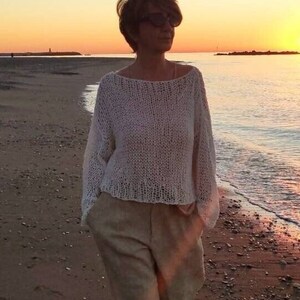 Pu&ograve; includere: Una donna cammina lungo una spiaggia sabbiosa al tramonto indossando un maglione bianco lavorato a maglia e pantaloni beige. Il testo "Sweater pattern Patron de punto" &egrave; in basso nell'immagine.