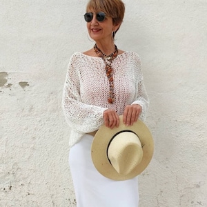 Pu&ograve; includere: Una donna indossa un maglione di crochet bianco, una gonna bianca e un cappello di paglia. Indossa una lunga collana con perline marroni e arancioni.