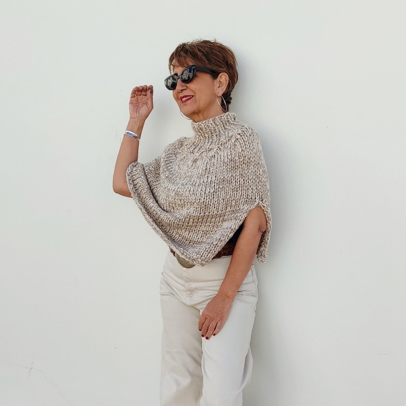 May include: A woman wearing a beige knitted poncho with a high neck and a brown belt. She is wearing white pants and black sunglasses.