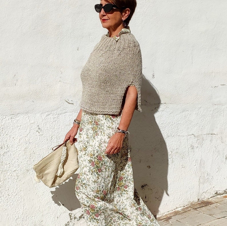 May include: A woman wearing a beige knitted cape with a floral print skirt and a beige bag.