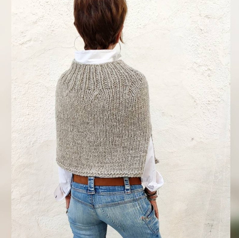 May include: A grey knitted poncho with a wide neckline and a brown leather belt. The poncho is made of a soft, textured yarn and has a relaxed fit.