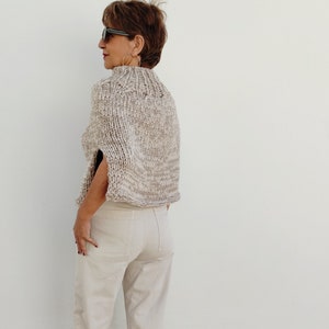 May include: A beige knitted cape with a high neck and short sleeves. The cape is made of a chunky yarn and has a slightly textured surface.