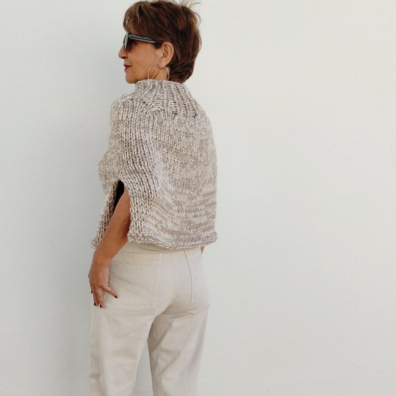 May include: A beige knitted cape with a high neck and short sleeves. The cape is made of a chunky yarn and has a slightly textured surface.