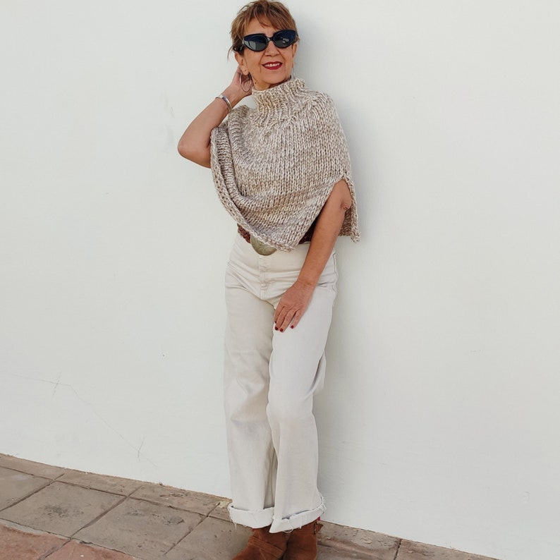May include: A woman wearing a beige knitted turtleneck poncho with a white background and a white pair of pants. She is wearing black sunglasses and has brown hair.