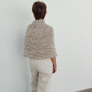May include: A beige knitted poncho with a high neck and a textured pattern. The poncho is worn by a person with short brown hair.