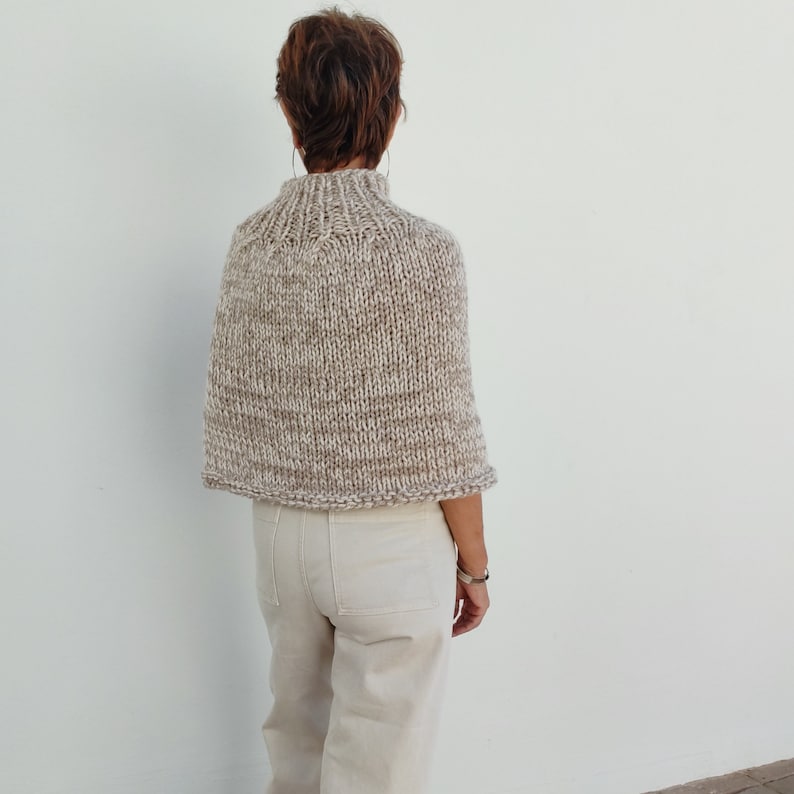May include: A beige knitted poncho with a high neck and a textured pattern. The poncho is worn by a person with short brown hair.
