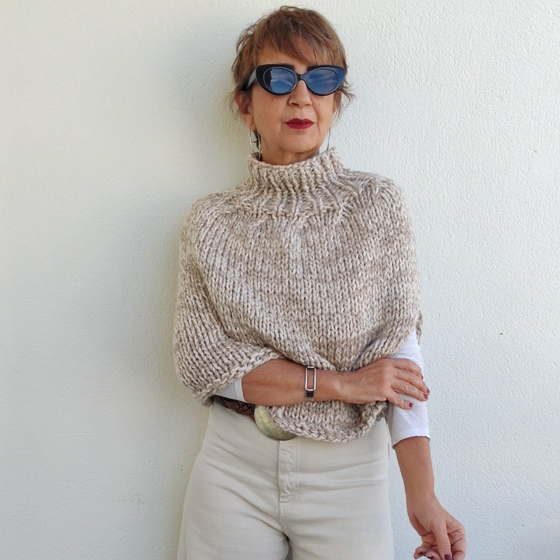 May include: A beige knitted poncho with a high neck and a subtle speckled pattern. The poncho is worn over a white shirt and beige pants.