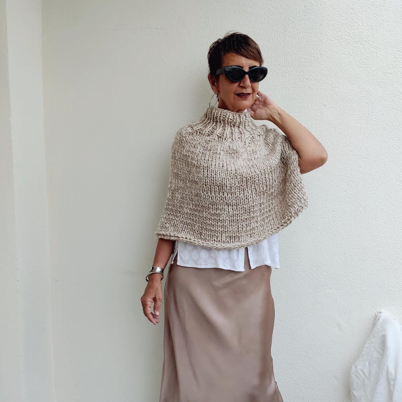 May include: A beige knitted poncho with a high neck and a loose fit. The poncho is worn over a white shirt and a brown skirt.