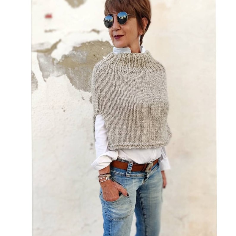 May include: A beige knitted cape with a high neck and short sleeves. The cape is made from chunky yarn and has a textured look.