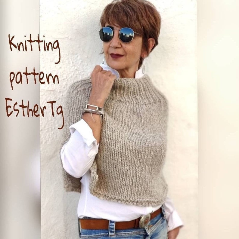 May include: A woman wearing a beige knitted poncho with a high neck and a white shirt underneath. The poncho has a simple design and is knitted in a chunky yarn. The woman is wearing jeans and a brown belt.