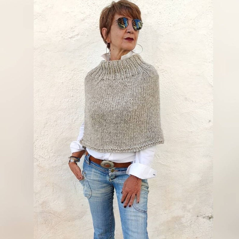 May include: A grey knitted poncho with a high neck and a loose fit. The poncho is made from a soft, textured yarn and has a subtle flecked pattern.