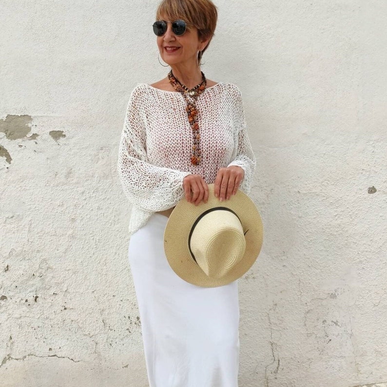 Pu&ograve; includere: Una donna indossa un maglione di crochet bianco, una gonna bianca e un cappello di paglia. Indossa una lunga collana con perline marroni e arancioni.