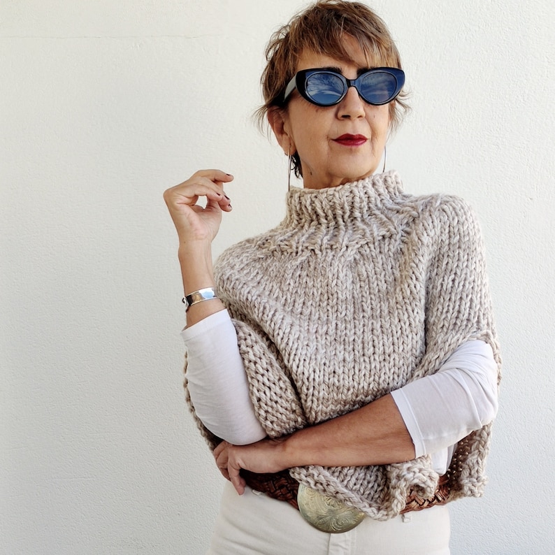May include: A beige knitted turtleneck sweater with a loose fit and a wide, flowing neckline. The sweater is made of a soft, textured yarn and has a casual, relaxed style.