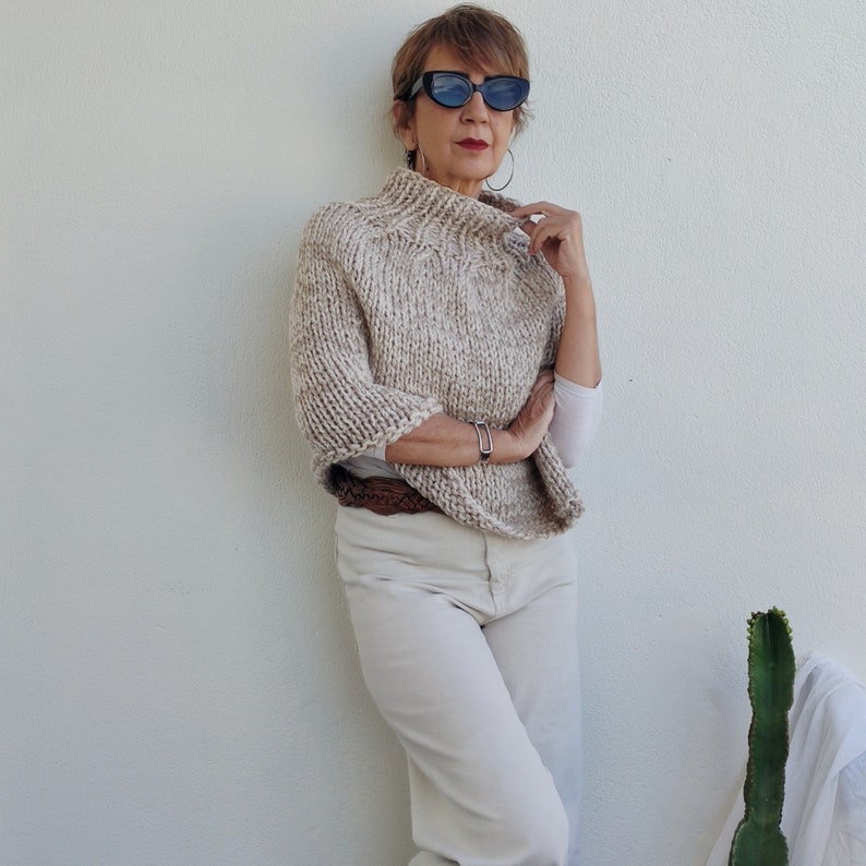 May include: A woman wearing a beige knitted poncho with a high neck and a brown leather belt. She is wearing white pants and black sunglasses.