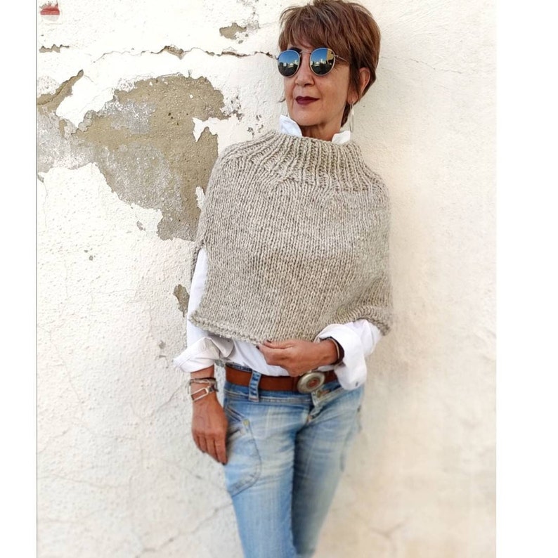May include: A woman wearing a beige knitted poncho with a high neck and a white shirt. She is wearing blue jeans and sunglasses.