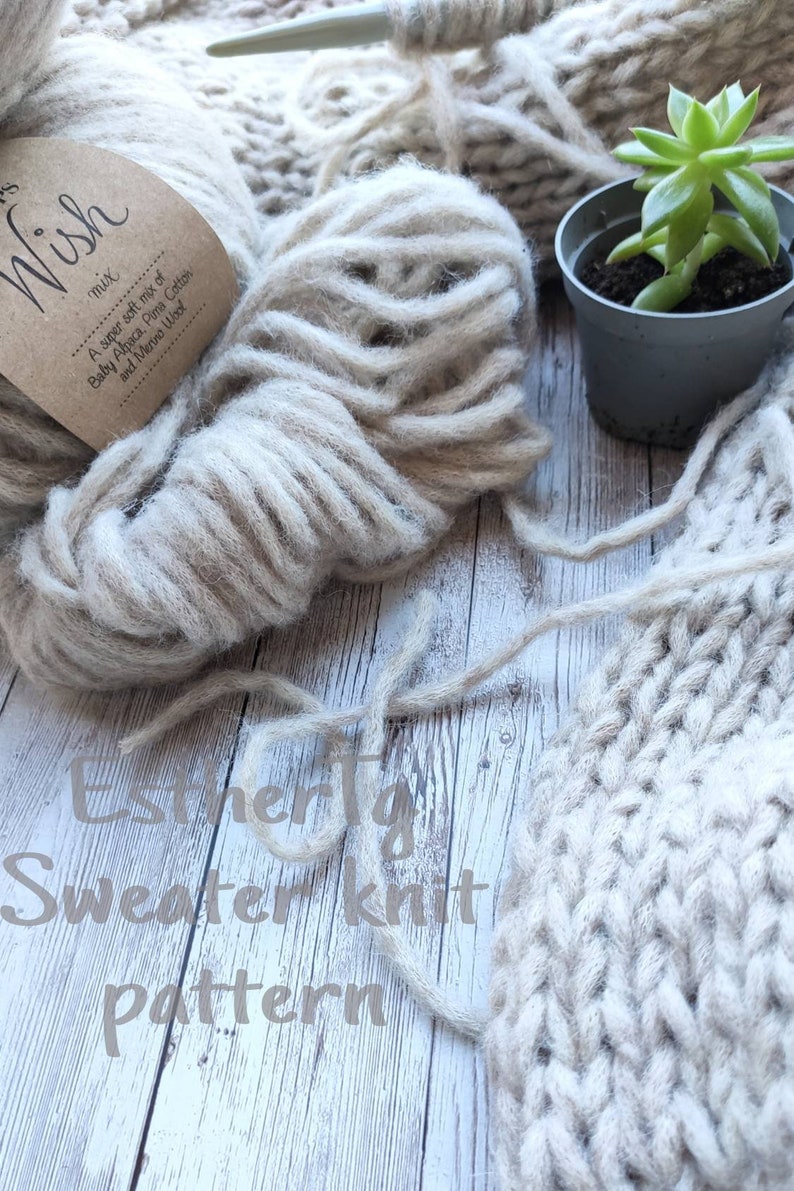 May include: A close-up of a ball of beige yarn with a brown label that reads "A super soft mix of Baby Alpaca, Pima Cotton and Merino Wool". The yarn is next to a knitted sweater made with the same yarn. The text "Estherty Sweater Knit Pattern" is written on the wooden surface.