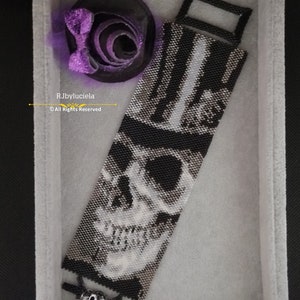 Può includere: Un braccialetto di perline bianco e nero con un design a forma di teschio e un cappello a cilindro. Il braccialetto ha una chiusura argentata ed è esposto in una scatola di velluto bianco. Il testo "RJbyluciela © All Rights Reserved" è visibile nell'immagine.