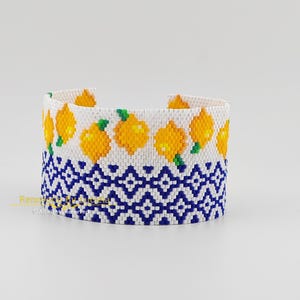 Può includere: Un braccialetto di perline con sfondo bianco decorato con limoni gialli e foglie verdi. La parte inferiore presenta un motivo geometrico blu e bianco. Il braccialetto è largo circa 5 cm.