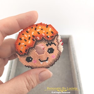 May include: A beaded donut-shaped brooch with a smiling face, featuring orange, brown, and peach beads. The brooch has a hole in the center and is held by a hand. A daisy and ladybug pin are in the background. Text on the image reads "Renovatio By Luciela © All Rights Reserved."